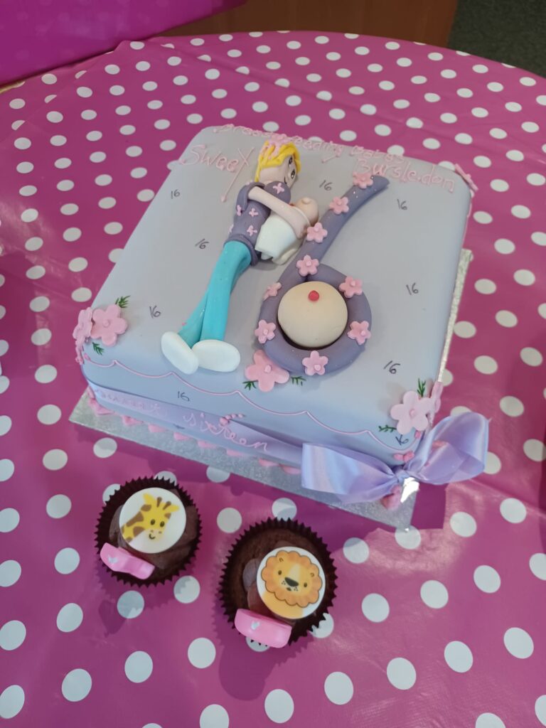 A square cake with the number 16 in fondant icing - the 1 is a woman with blonde hair lying down with a breastfeeding baby lying next to her. The 6 has a boob in the centre. The background is a pink table cloth with white polka dots.